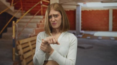 İnşaat alanındaki genç sarışın kadın, bileğini inceliyor bitmemiş duvarlar ve iskelelerle çevrili..