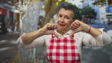 Önlüklü, gri saçlı yaşlı bir kadın. İki başparmağı aşağıda, kalabalık bir caddede ayakta duruyor..