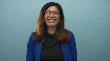 Orta yaşlı, gözlüklü, açık mavi duvarlı bir kadın stüdyoda kamerayı işaret ediyor..