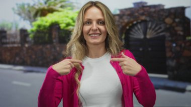 Pembe kazaklı bir kadın arka planda yeşil yapraklarla güneşli bir caddede kendini işaret ederek gülümsüyor açık hava canlılığıyla kişisel güvenini harmanlıyor..