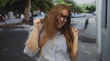 Güneş ışığında park halindeki arabaların arasında dans ederken gözlük takan beyaz bir kadın yumruk kaldırıyor..