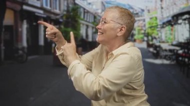 Kadın, diğer el, kısa saç ve gözlük ile gülümseyerek ve göğsüne dokunarak yaya caddesinde ileri doğru işaret eder; neşeli neşe.