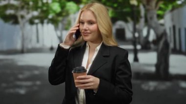 Siyah takım elbiseli genç bir kadın telefonda konuşuyor. Elinde kahve fincanı var. Şehir caddesinde ağaçlar ve bulanık binalar..