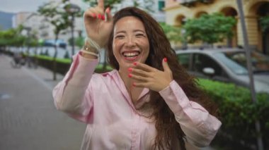 Güneş ışığı altındaki caddede, kırmızı ojeli tırnaklar gösteren ve açık bir gülümsemeyle gülen bir kadın..