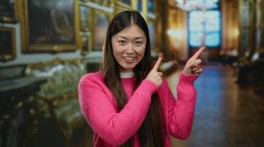 Pembe kazaklı bir kadın müzenin içinde gülümsüyor ve işaret ediyor. Sanat dolu duvarlar ve tarihi dekorlar sergiliyor. Kapalı bir kültür ortamında neşe ve zarafet anı yakalıyor..