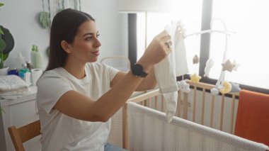 Genç bir kadın, bebek tulumunu bina odasındaki asılı bir cep telefonunun yanında ahşap bir beşikte tutarken gülümsüyor. Annelik şefkati..