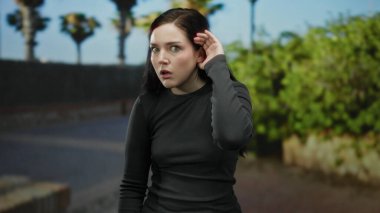 Sokakta gezen kadın şaşırmış görünüyor. Elleri havada, güneşli bir arka planda, güneşli bir günde ağaçlar ve şehir manzarası eşliğinde günlük siyah kıyafetler giyiyor..