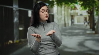 Kumral saçlı, gözlüklü bir kadın, güneşli bir sokakta sorgu jesti olarak ellerini uzatıyor. Etrafı bulanık yeşillik ve mimariyle çevrili. Merakını dile getiriyor..