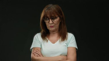 Gözlüklü yaşlı bir kadın siyah bir arka plana karşı duruyor. Kollarını kavuşturup izole edilmiş bir stüdyoda düşünceli bir ifade sunuyor..