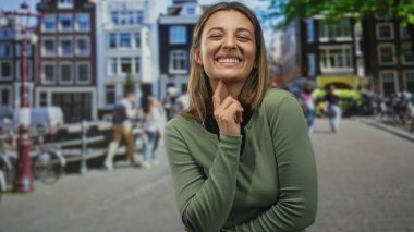 Canlı kaldırımlı bir sokakta parmakları yanaklarına değen, omuzları rahat, neşeli bir şekilde gülümseyen bir kadın..