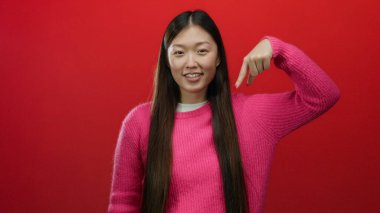 Pembe süveter giyen, uzun siyah saçlı, koyu kırmızı arka planda gülümseyen, aşağıya bakan, neşeli bir ifade ve canlı bir jest tasvir eden bir kadın..