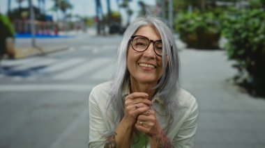 Yaşlı, kır saçlı, dövmeli bir kadın güneşli bir caddede sıcacık gülümsüyor, kaldırımda ağaçların oluşturduğu bir şehir ortamında neşe ve pozitiflik sergiliyor..