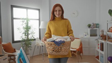 Çamaşır sepetiyle gülümseyen bir kadın, parlak pencereli ve dekorlu, ev temizlik rutinini sergiliyor..