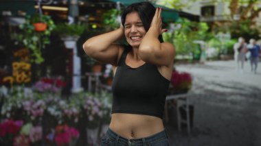 Kadın elleriyle kulaklarını kapatıyor bir sokak çiçeğinin önünde güneş ışığı altında saksı çiçeklerinin yanında dururken; hüsran..