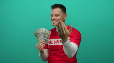 Amerikan dolarlarını elinde kırmızı bir gömlek içinde tutan genç adam yeşil arka planda gönüllü bir metin servetini ve cömertliğini neşeyle işaret ediyor ve işaret ediyor..