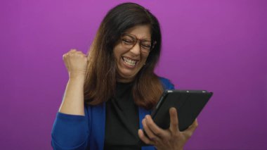 Gülümseyen orta yaşlı İspanyol kadın mor stüdyoda tablet tutuyor ve tıkırdatıyor; heyecan güçlendiriyor.