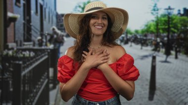 Hasır şapkalı ve kırmızı omuzlu bir kadın, elleri göğsünde ve güneşli kaldırımlı bir caddede parmaklıklar ve park edilmiş bisikletlerle gülümsüyor. Minnettarlık..