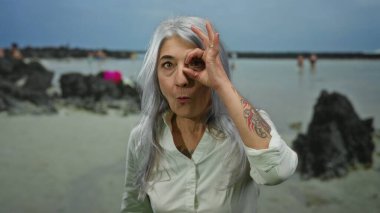 Üst düzey bir kadın, kır saçlı ve neşeli bir ifadeyle manzaralı bir sahil sahilinde dikilip açık havanın tadını çıkarırken, gözünün önünde iyi bir jest yapıyor.