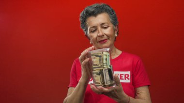 Gri saçlı yaşlı kadın para kavanozunu tutarken canlı kırmızı bir zemin için gönüllü oluyor, yardım ve finansal destek temaları sergiliyor..