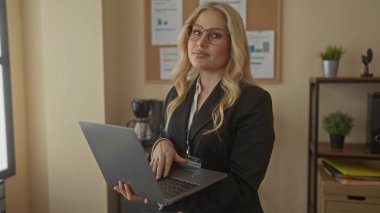 Ofiste dizüstü bilgisayar kullanan genç bir kadın. Gözlük takıp işe odaklanıyor. Arkaplanda çizelgeler var.