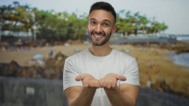 Elleri kameraya doğru uzanmış, kumsalda gülümseyen genç İspanyol adam, rahat ve tatil havasıyla açık deniz manzarasında neşe yakalıyor..