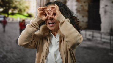 Kıvırcık saçlı, olgun bir İspanyol kadın güneşli bir caddede dürbünle el kol hareketi yapar..