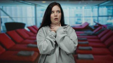 Kaplıcada siyah saçlı bir kadın rahatsızlığını dile getiriyor. Sağlık merkezinde boğazına dokunuyor. Arka planda kırmızı sandalyeler var..