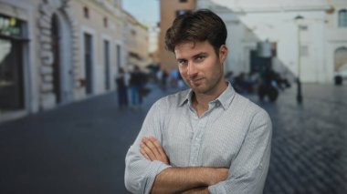 Kolları çaprazlanmış bir adam kaldırım taşı caddesinde sıradan çizgili bir düğme gömlekle kaldırımları ve binaları bulanık gösteren bir adam..