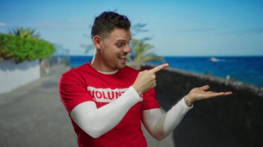 Kırmızı gömlekli, gülümseyen ve arkasında okyanus manzarası olan genç bir adam. Parlak gökyüzü boyunca geziniyor. Neşeli ve davetkar bir deniz manzarası sergiliyor.