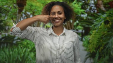 Beyaz çizgili gömlekli kadın, yeşil yapraklarla kaplı parkta burun buruna, rahat duruş ve dostça ifadeyle gülümseyen;.