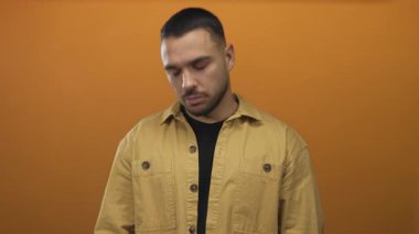 Sarı ceketli adam, turuncu stüdyoda yüzünü gösteren tarafsız bir ifadeyle aşağıya bakıyor..