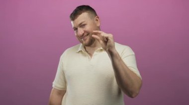 Pembe stüdyoda krem polo giyerken az miktarda parmak çimdikleyen bir adam, şakacı bir şaka yapıyor..