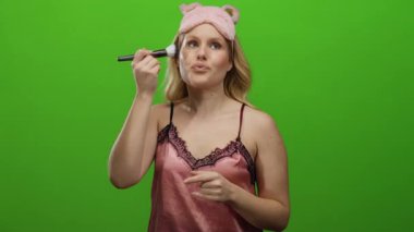 Pembe gecelikli bir kadın yeşil arka planda fırçayla makyaj yaparken sevimli bir uyku maskesi takıyor..