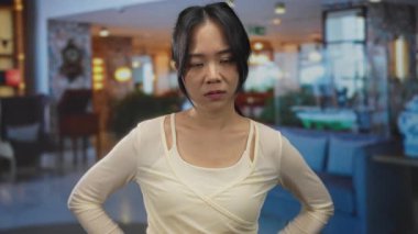 Bir kadın otel lobisinde durmuş modern dekorun ortasında ciddi bir ifade sergiliyor. Zarif bir ortamda sakin ve düşünceli bir ruh halini yansıtıyor..