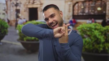 İşlek şehir caddesinde parmaklarıyla hashtag jesti oluşturan genç İspanyol adam; neşeli sosyal trend.