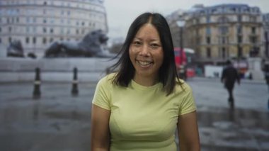 Elle yanağa gülümseyen kadın ve çapraz kollar, sarı gömlek ve görünür yüzük, taş çeşme ve binalar yanında sokakta; dinginlik güven.