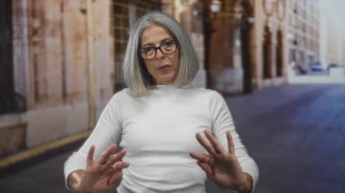 Orta yaşlı, gri saçlı, gözlüklü, beyaz saçlı bir kadın avuçlarını kaldırır ve iki elini de kaldırır..