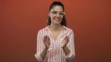 Turuncu stüdyoda gülümseyen ve el çırpan bir kadın, gözlük takan ve kolları kıvrılmış pembe ve beyaz çizgili bir gömlek giyen; sevinç kutlaması.