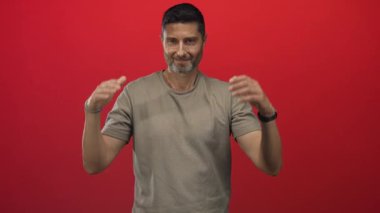 Kırmızı stüdyoda elleri havada gülümseyen, bej tişört, saat ve bilezik giyen, avuç içi açık kameraya bakan ve rahat duruşu olan bir adam; dostça karşılama.