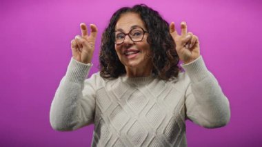 Kıvırcık saçlı, olgun bir İspanyol kadın mor arka planda zafer jesti yaparak neşe ve pozitiflik gösteriyor..