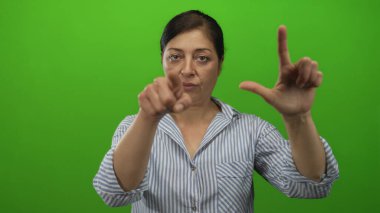 Orta yaşlı İspanyol kadın yeşil stüdyo ortamında parmağı ve işaret parmağıyla ezik el işareti yapıyor..