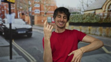 Kırmızı tişört giyen bir adam işaret parmağını şehir sokaklarında yukarı kaldırıyor; mutluluk güven ifadesi.