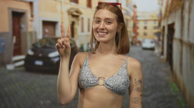 Üzerinde barış işareti olan bikinili genç sarışın kadın renkli binalarla çevrili bir şehir sokağında duruyor, kaygısız bir yaz havasını somutlaştırıyor..