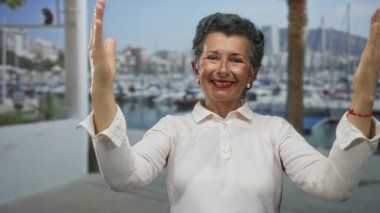 Gri saçlı yaşlı bir kadın, büyük elleri olan, deniz kıyısındaki limanda neşeyle gülümseyen, arka planda tekneleri olan, mutluluğu somutlaştıran..