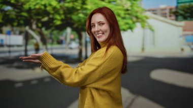 Güneşli bir şehir caddesinde, ağaçlar ve binalarla dolu sarı bir kazakla gülümseyen bir kadın, şehir atmosferiyle neşeli bir açık hava sahnesi yaratıyor..