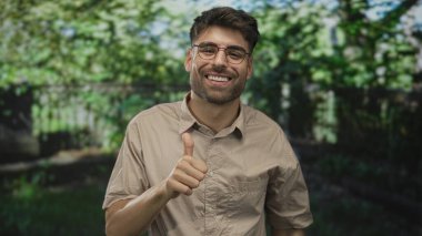 Genç İspanyol adam yeşil orman ortamında gülümsüyor ve başparmak işareti yapıyor; mutluluk motivasyon güveni.