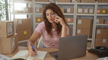 Telefonu kulağına tutan ve kalemle yazan kadın depolama raflarında istiflenmiş paketlerin yanında; küçük işletme stresi.