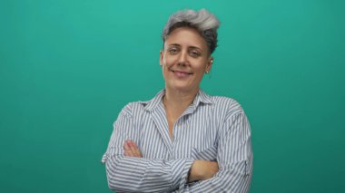 Kısa saçlı ve sakin yüzlü bir kadın çizgili bir gömlek giyiyor ve teal stüdyo ortamında kollarını kavuşturuyor. Reddedildi..