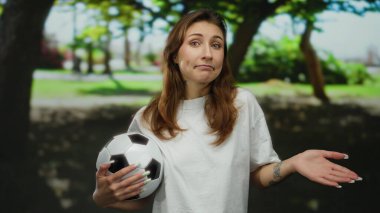 Güneşli park ortamında elinde futbol topu tutan bir kadın yemyeşil bir ortamda neşeli bir ifade sergiliyor. Dışarıda kaygısız bir boş zamanı yansıtıyor..