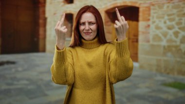 Üniversite kampüsünün dışında el kol hareketi yapan, tarihsel eğitim ortamında meydan okuyan, hardal kazak giyen, kızıl saçlı güzel bir kadın..